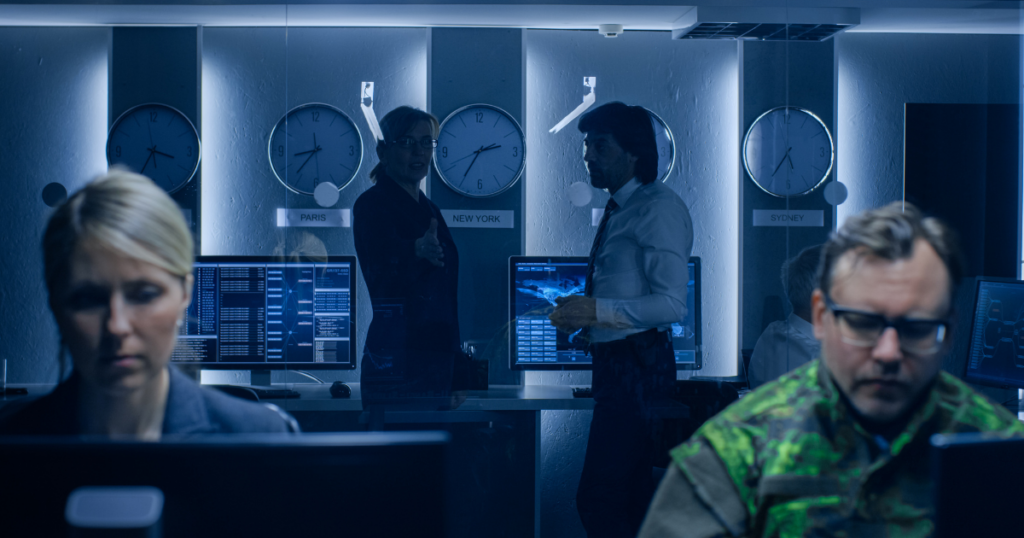 Image of a dark office, two leadership team members standing in the back of the room, and two other employees working on their computers.